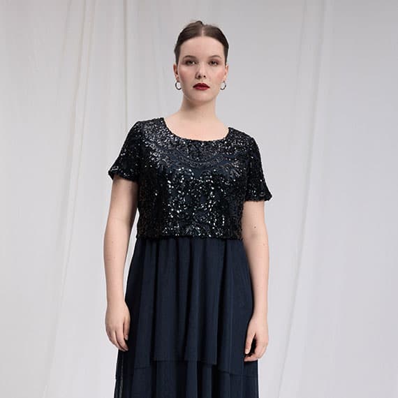 Frau trägt ein langes schwarzes Kleid mit Pailletten-Oberteil und kurzen Ärmeln vor weißem Hintergrund