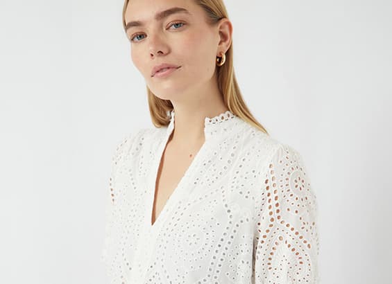 Frau trägt weiße Bluse mit Lochmuster und Stehkragen, dazu goldene Ohrringe.