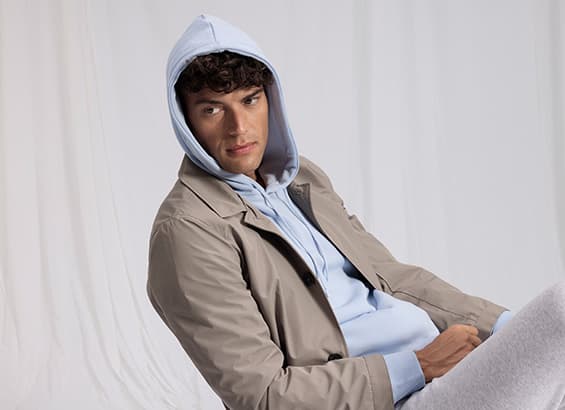 Mann sitzt entspannt, trägt hellblauen Hoodie und beigefarbene Jacke. Gesicht unscharf. Hintergrund weiß.