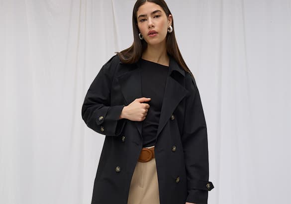 Frau trägt schwarzen Mantel, schwarzes Oberteil und beige Hose mit Gürtel vor weißem Hintergrund