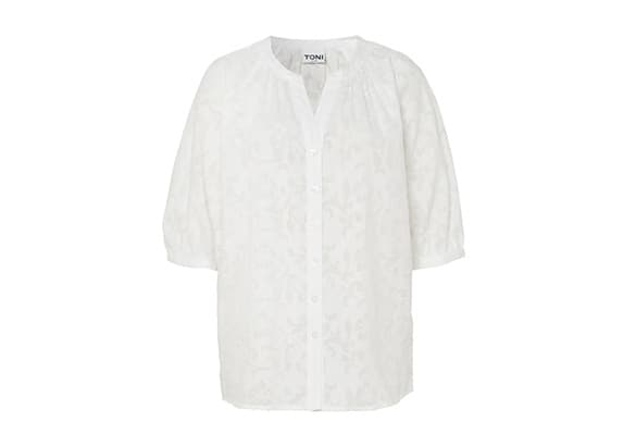 Femme portant un trench-coat noir et un pantalon beige, posant devant un fond blanc.